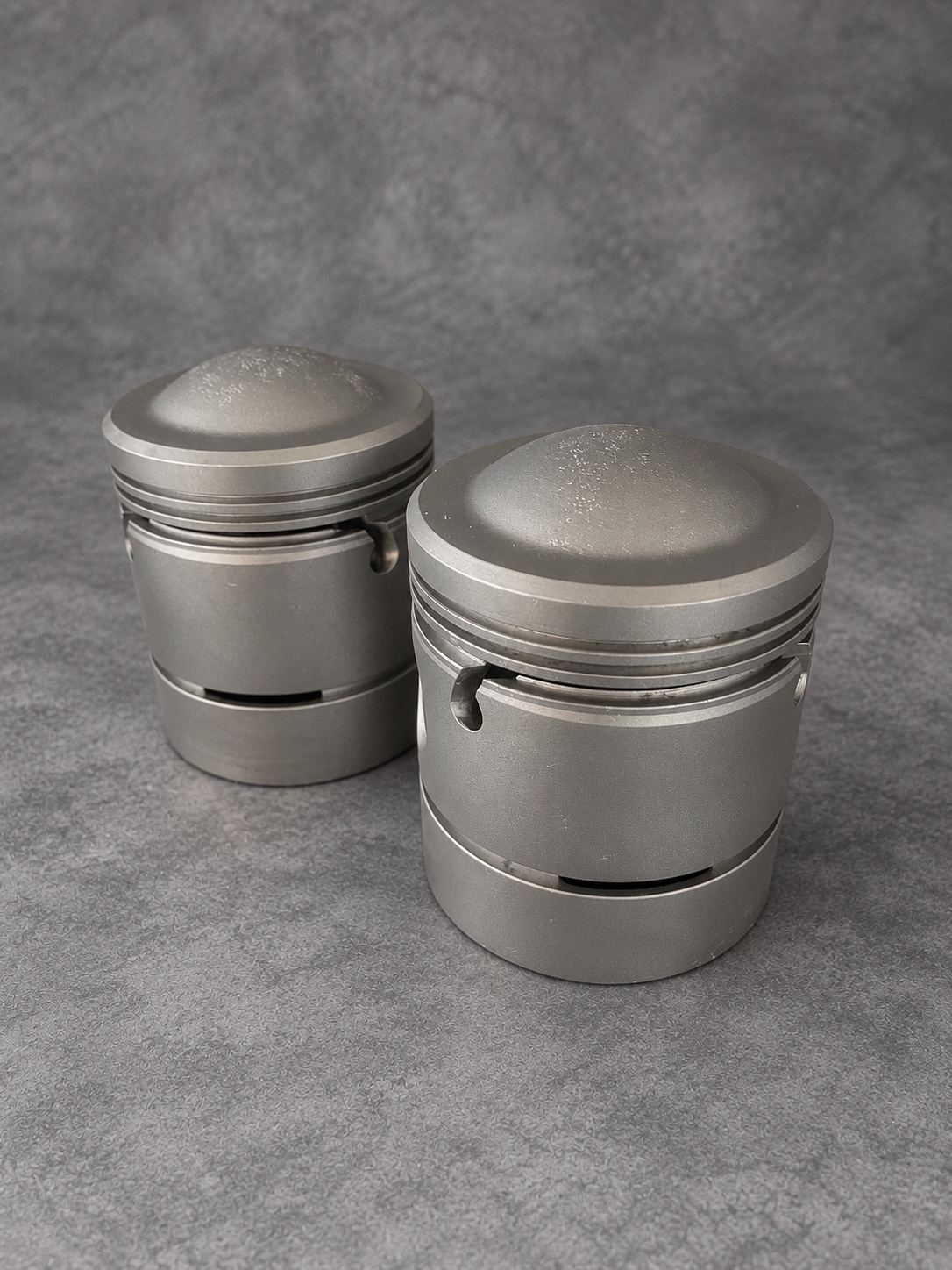 Two vapour-blasted pistons standing upright, showing domed crowns and ring grooves